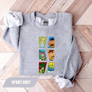 May include: A sport grey crewneck sweatshirt featuring a graphic print of six Toy Story characters arranged in two columns. The characters include Bo Peep, Mr. Potato Head, Buzz Lightyear, Woody, Rex, and an alien, with the text 30 YEARS below.