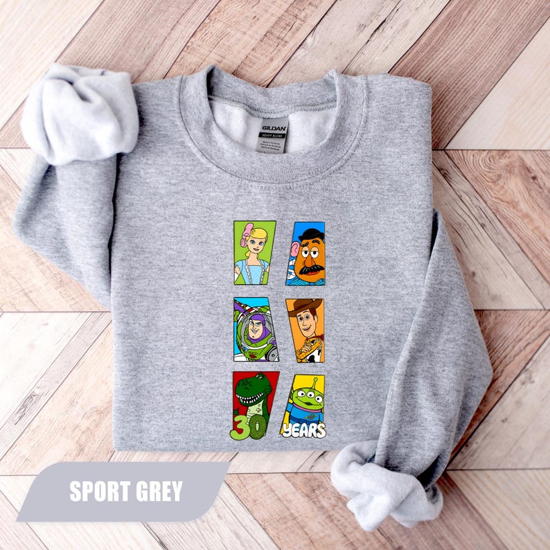 May include: A sport grey crewneck sweatshirt featuring a graphic print of six Toy Story characters arranged in two columns. The characters include Bo Peep, Mr. Potato Head, Buzz Lightyear, Woody, Rex, and an alien, with the text 30 YEARS below.