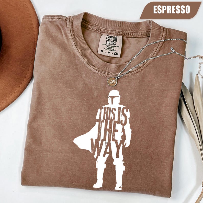 May include: An espresso-colored t-shirt featuring a white graphic of a helmeted character silhouette with the text "THIS IS THE WAY" integrated into the design. The shirt has a crew neckline and a Comfort Colors brand label inside the collar.