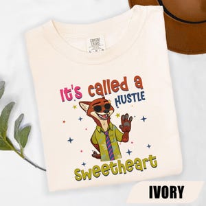 K&ouml;nnte beinhalten: Elfenbeinfarbenes T-Shirt mit dem Text "It's called a HUSTLE sweetheart" und einem Cartoon-Fuchs mit Sonnenbrille, gr&uuml;nem Hemd und Krawatte. Das Shirt hat auch Sterne. Das Shirt ist mit "IVORY" gekennzeichnet.