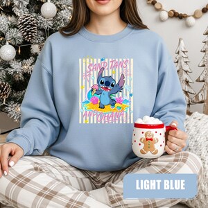 May include: A light blue crewneck sweatshirt featuring a graphic of the character Stitch on a beach with the text SAND, TANS AND OHANA. The design includes a sun, palm trees, seashells, and a starfish. A white mug with a gingerbread man print is held.