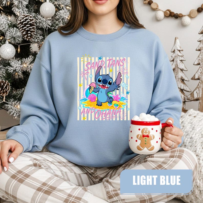 May include: A light blue crewneck sweatshirt featuring a graphic of the character Stitch on a beach with the text SAND, TANS AND OHANA. The design includes a sun, palm trees, seashells, and a starfish. A white mug with a gingerbread man print is held.