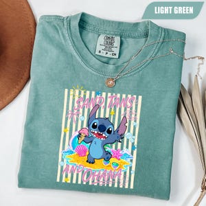 Op de afbeelding: Een lichtgroen T-shirt met korte mouwen en een afbeelding van het personage Stitch met een ijshoorntje. Het ontwerp bevat verticale strepen, een zon, palmbomen, schelpen en de tekst "SAND, TANS AND OHANA" in roze letters.