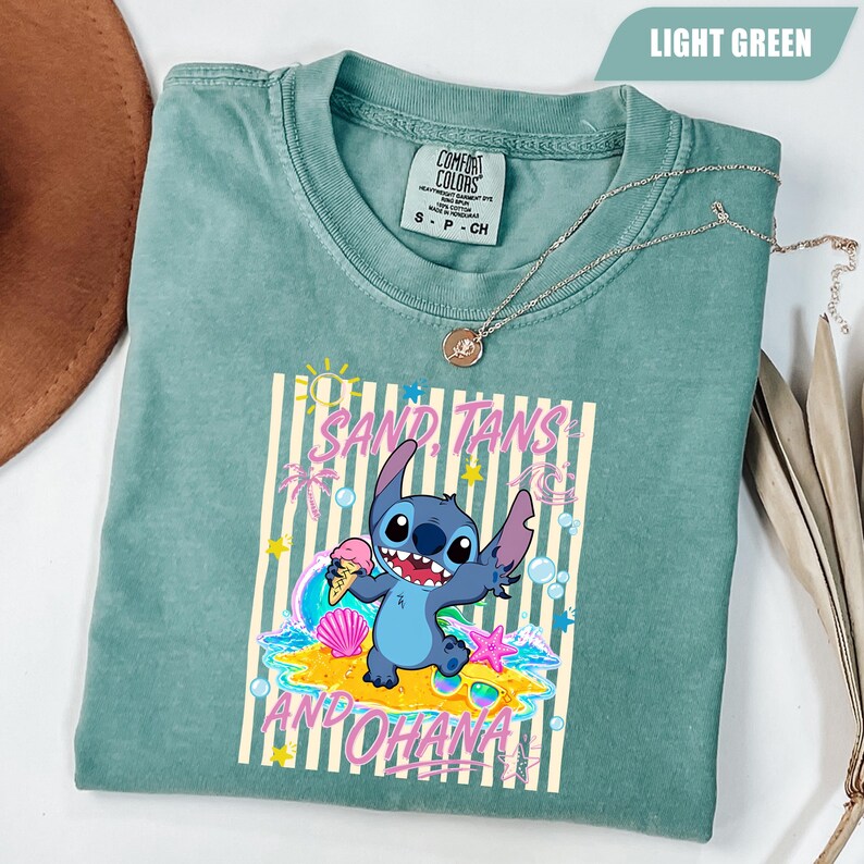 Op de afbeelding: Een lichtgroen T-shirt met korte mouwen en een afbeelding van het personage Stitch met een ijshoorntje. Het ontwerp bevat verticale strepen, een zon, palmbomen, schelpen en de tekst "SAND, TANS AND OHANA" in roze letters.