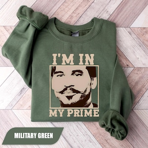 Puede incluir: Sudadera verde militar con un gráfico de la cara de un hombre y el texto "I'M IN MY PRIME". La sudadera tiene cuello redondo y está hecha de un material suave. El texto "MILITARY GREEN" está en la parte inferior.
