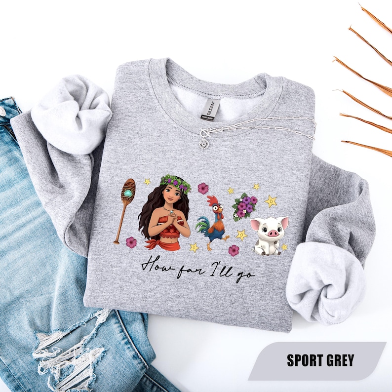 Pu&ograve; includere: Felpa grigio sport con una grafica colorata di una donna polinesiana, un gallo e un maiale, con la scritta "How far I'll go". La felpa ha un girocollo e maniche lunghe. L'immagine mostra anche un paio di jeans.