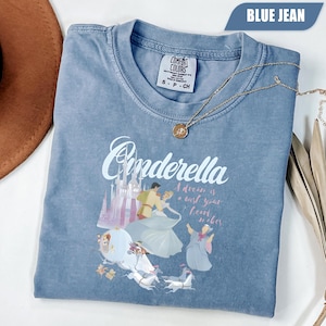 Peut inclure: Un t-shirt bleu jean Comfort Colors avec un motif Cendrillon. Le motif comprend le mot "Cendrillon", un ch&acirc;teau et des personnages du film. Un collier en or avec un pendentif est drap&eacute; sur le t-shirt.