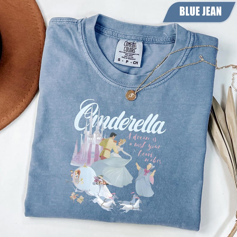 Peut inclure: Un t-shirt bleu jean Comfort Colors avec un motif Cendrillon. Le motif comprend le mot "Cendrillon", un ch&acirc;teau et des personnages du film. Un collier en or avec un pendentif est drap&eacute; sur le t-shirt.