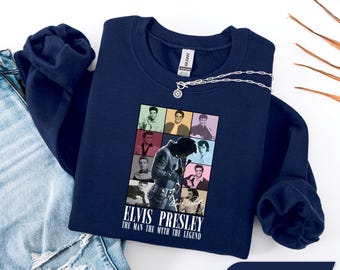 May include: Navy blue sweatshirt featuring a graphic of Elvis Presley in various poses. The graphic includes the text "ELVIS PRESLEY THE MAN THE MYTH THE LEGEND". A silver necklace is draped across the sweatshirt. The sweatshirt is folded on top of a pair of blue jeans.