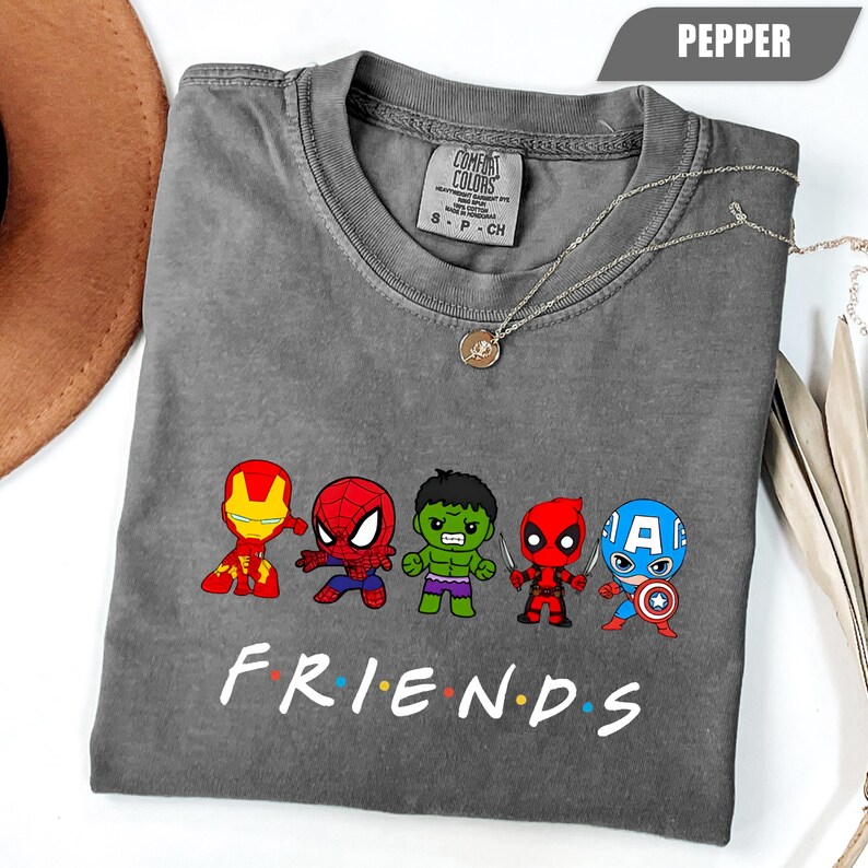 May include: A gray Comfort Colors t-shirt featuring cartoon versions of Marvel superheroes: Iron Man, Spider-Man, Hulk, Deadpool, and Captain America. The word "FRIENDS" is printed below the characters in white. A silver necklace is draped across the shirt.