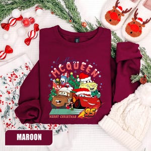 May include: Maroon sweatshirt featuring a graphic of Lightning McQueen and other Cars characters in Santa hats with the text "Merry Christmas". The sweatshirt is surrounded by holiday decorations, including candy canes and a white knit hat.