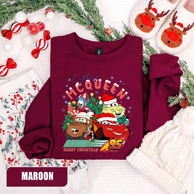 May include: Maroon sweatshirt featuring a graphic of Lightning McQueen and other Cars characters in Santa hats with the text "Merry Christmas". The sweatshirt is surrounded by holiday decorations, including candy canes and a white knit hat.