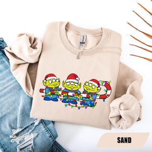 May include: A sand-colored sweatshirt featuring a graphic of three green aliens wearing Santa hats and holding a candy cane-like string of lights. The aliens are in blue outfits. The word "SAND" is at the bottom.