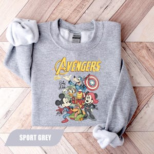 May include: A sport grey crewneck sweatshirt featuring a graphic of Disney characters dressed as Avengers. The print includes Mickey Mouse, Minnie Mouse, Goofy, Donald Duck, and Pluto with the text The Avengers and character signatures.