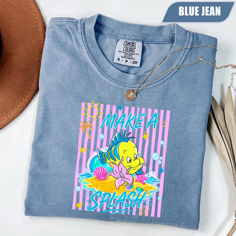 May include: Blue jean colored t-shirt with a graphic of a yellow fish, pink seashell, and the words "MAKE A SPLASH". The shirt has a crew neck and short sleeves. A gold necklace is draped across the shirt.