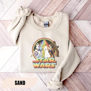 May include: Sand-colored sweatshirt featuring a Star Wars graphic. The graphic includes characters like Luke Skywalker, Princess Leia, and Chewbacca, set within a rainbow circle. The text "STAR WARS" is displayed below the characters in a colorful, retro font.