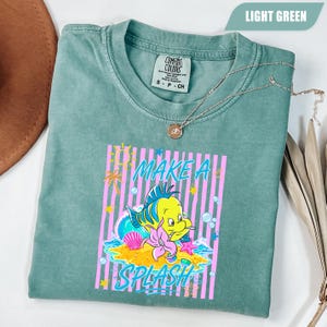 May include: A light green t-shirt with a graphic of a yellow fish with blue and green accents, a pink starfish, and the words "MAKE A SPLASH" in a colorful design. The shirt is folded and has a silver necklace on top.