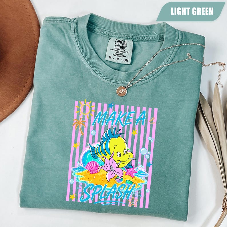 May include: A light green t-shirt with a graphic of a yellow fish with blue and green accents, a pink starfish, and the words "MAKE A SPLASH" in a colorful design. The shirt is folded and has a silver necklace on top.