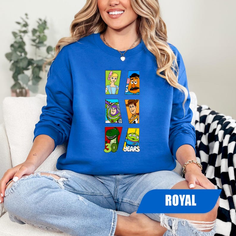 May include: A royal blue long-sleeve sweatshirt featuring a graphic print of six Toy Story characters in individual rectangular frames, including Bo Peep, Mr. Potato Head, Buzz Lightyear, Woody, Rex, and an alien, with the text 30 YEARS.