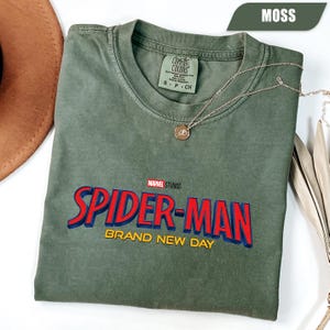 Może przedstawiać: Zielona koszulka w kolorze mchu z napisem "SPIDER-MAN BRAND NEW DAY" w kolorze czerwonym i niebieskim. Koszulka ma okrągły dekolt i kr&oacute;tkie rękawy. Złoty naszyjnik z wisiorkiem jest zarzucony na koszulkę. Koszulka jest wykonana z miękkiego, wygodnego materiału.
