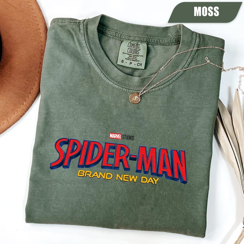 Może przedstawiać: Zielona koszulka w kolorze mchu z napisem "SPIDER-MAN BRAND NEW DAY" w kolorze czerwonym i niebieskim. Koszulka ma okrągły dekolt i kr&oacute;tkie rękawy. Złoty naszyjnik z wisiorkiem jest zarzucony na koszulkę. Koszulka jest wykonana z miękkiego, wygodnego materiału.