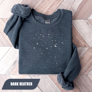 May include: Dark heather grey sweatshirt with a celestial design of stars and a crescent moon. The text "DARK HEATHER" is displayed on a dark grey banner. The sweatshirt is folded on a light brown wooden surface.