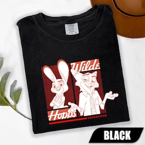 Puede incluir: Camiseta negra con una ilustraci&oacute;n de dibujos animados de Judy Hopps y Nick Wilde de Zootopia. El dise&ntilde;o incluye las palabras "Hopps" y "Wilde" en rojo y blanco. Fabricado por Comfort Colors.