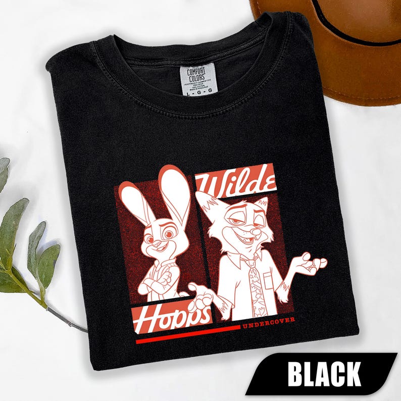 Puede incluir: Camiseta negra con una ilustraci&oacute;n de dibujos animados de Judy Hopps y Nick Wilde de Zootopia. El dise&ntilde;o incluye las palabras "Hopps" y "Wilde" en rojo y blanco. Fabricado por Comfort Colors.