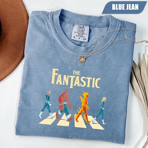 Peut inclure: Un t-shirt bleu jean avec un motif représentant quatre personnages marchant sur un passage piéton et le texte THE FANTASTIC imprimé au-dessus. Le t-shirt possède un col rond et une étiquette de marque Comfort Colors à l'intérieur du col.