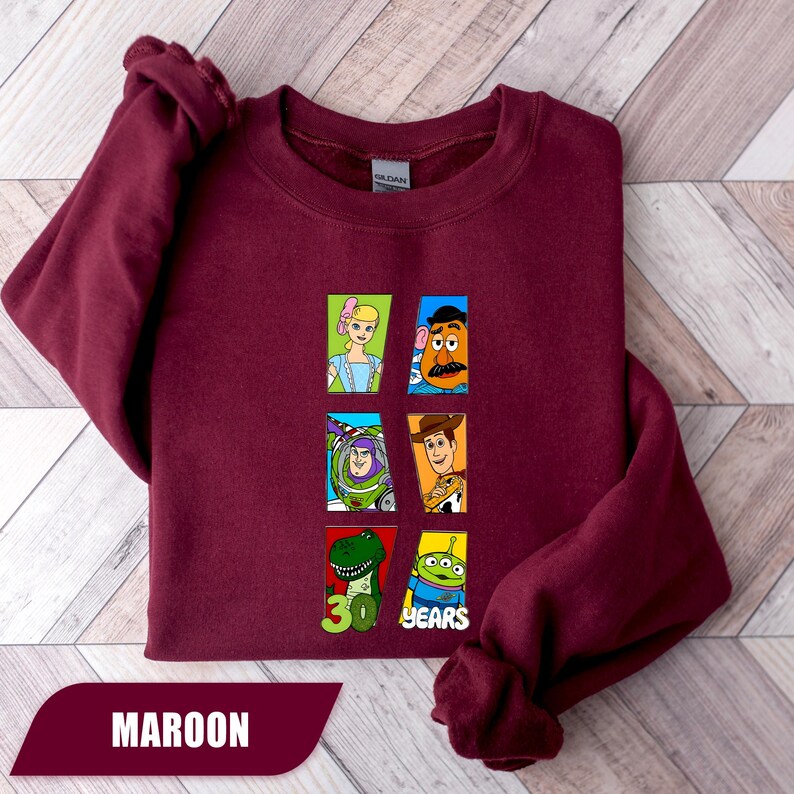 May include: A maroon crewneck sweatshirt featuring a graphic print of six Toy Story characters arranged in two columns. The design includes Bo Peep, Mr. Potato Head, Buzz Lightyear, Woody, Rex, and an alien, with the text 30 YEARS printed below.