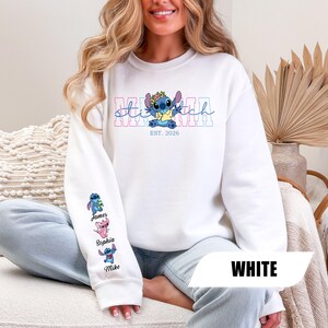 May include: A white long-sleeve sweatshirt featuring a graphic of the character Stitch wearing a flower crown, with the text MAMA and EST. 2026. The right sleeve displays three small character illustrations with the names James, Sophia, and Mike.