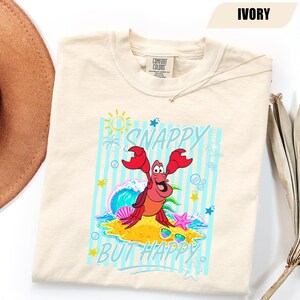 May include: Ivory-colored t-shirt featuring a cartoon crab with the text "SNAPPY BUT HAPPY" in a turquoise font. The crab is red and orange, with a beach scene background including a wave, sun, and sunglasses.