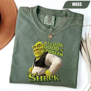 Pode incluir: Camiseta verde musgo com um gráfico de Shrek e o texto "I look good in green" e o nome "Shrek". A camiseta tem gola redonda e mangas curtas. Um colar de ouro está sobre a camiseta.