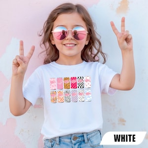 May include: A white short-sleeved t-shirt featuring a graphic print of twelve soda cans arranged in two rows. Each can displays the Dr. Pepper logo and a unique pattern, including leopard print, checkerboard, zebra stripes, cow print, and floral designs.
