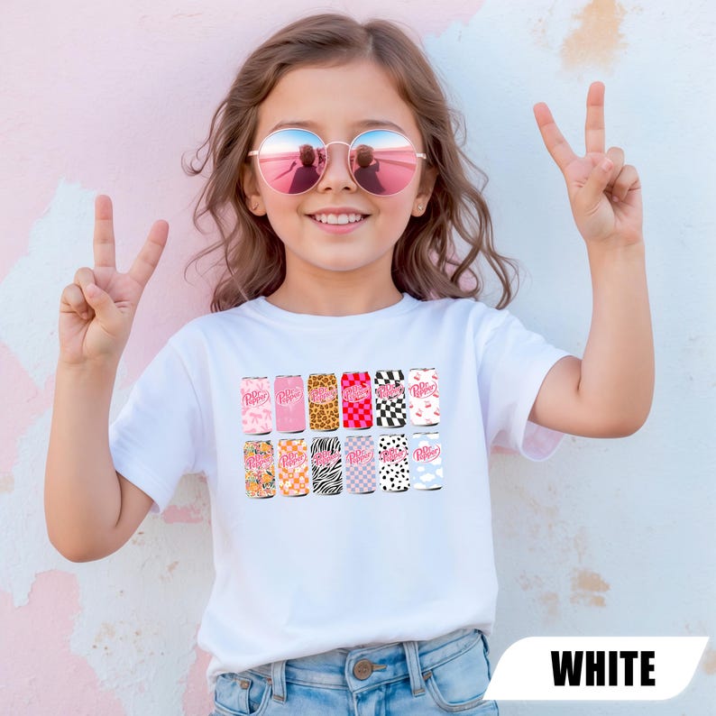 May include: A white short-sleeved t-shirt featuring a graphic print of twelve soda cans arranged in two rows. Each can displays the Dr. Pepper logo and a unique pattern, including leopard print, checkerboard, zebra stripes, cow print, and floral designs.