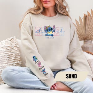 May include: A sand-colored crewneck sweatshirt featuring a graphic of the character Stitch wearing a flower crown. The text reads "stitch" and "EST. 2026". The sleeve displays three small character illustrations with the names James, Sophia, and Mike.