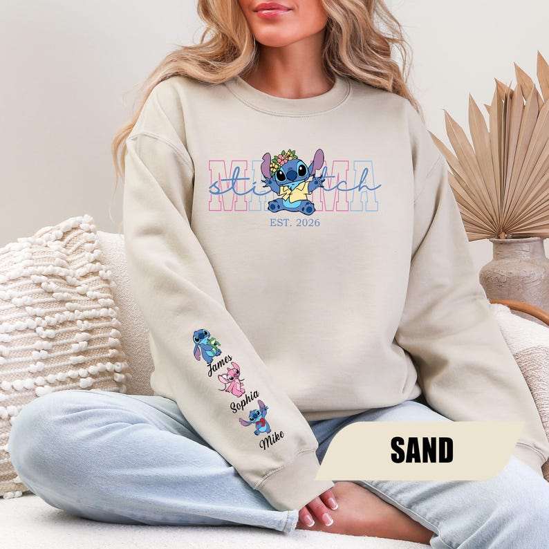 May include: A sand-colored crewneck sweatshirt featuring a graphic of the character Stitch wearing a flower crown. The text reads "stitch" and "EST. 2026". The sleeve displays three small character illustrations with the names James, Sophia, and Mike.
