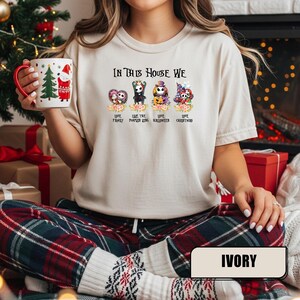 May include: Ivory-colored t-shirt with the text "IN THIS HOUSE WE LOVE FAMILY, LIKE THE PUMPKIN KING, LOVE HALLOWEEN, LOVE CHRISTMAS." The shirt features cartoon characters. The person is wearing plaid pants and holding a mug.