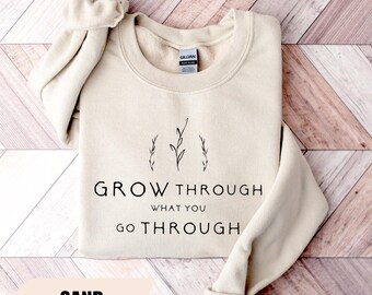 Può includere: Una felpa color sabbia con la scritta nera "GROW THROUGH WHAT YOU GO THROUGH" e illustrazioni minimaliste di piante. La parola "SAND" è in basso. La felpa è piegata su una superficie di legno.