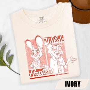 Puede incluir: Camiseta de color marfil con un gr&aacute;fico rojo de los personajes de dibujos animados Judy Hopps y Nick Wilde de Zootopia. Los textos "Wilds" y "Hopps" tambi&eacute;n son rojos. La camiseta es de material suave.
