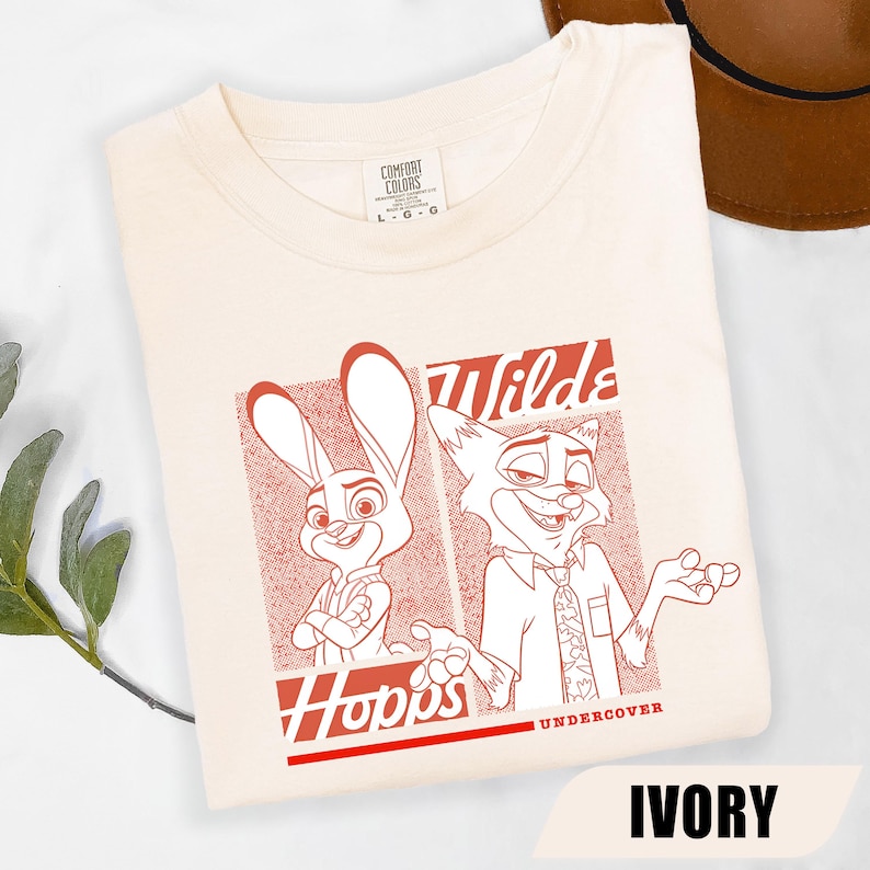 Puede incluir: Camiseta de color marfil con un gr&aacute;fico rojo de los personajes de dibujos animados Judy Hopps y Nick Wilde de Zootopia. Los textos "Wilds" y "Hopps" tambi&eacute;n son rojos. La camiseta es de material suave.