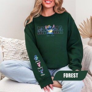 May include: A forest green crewneck sweatshirt featuring a graphic of the character Stitch wearing a flower crown above the text MAMA and EST. 2026. The left sleeve displays three small character figures with the names James, Sophia, and Mike.