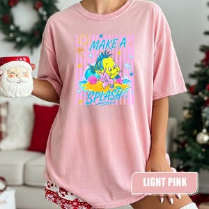May include: Light pink t-shirt featuring a cartoon illustration of a yellow fish with blue and green accents, a seashell, and the words "MAKE A SPLASH". The shirt has a relaxed fit and short sleeves.