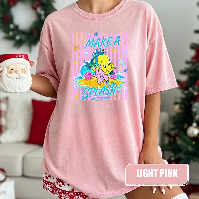 May include: Light pink t-shirt featuring a cartoon illustration of a yellow fish with blue and green accents, a seashell, and the words "MAKE A SPLASH". The shirt has a relaxed fit and short sleeves.