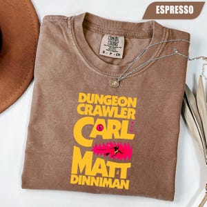 May include: Espresso-coloured t-shirt with the text "Dungeon Crawler Carl" in yellow and pink. The shirt features a graphic of a figure in a pink background. A silver necklace with a pendant is draped across the shirt.