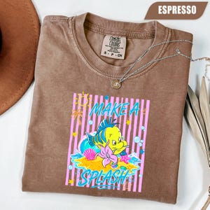May include: Espresso-colored t-shirt with a graphic of a yellow and blue cartoon fish with the text "MAKE A SPLASH". The design includes pink stripes, a seashell, and a starfish. A silver necklace is draped across the shirt.