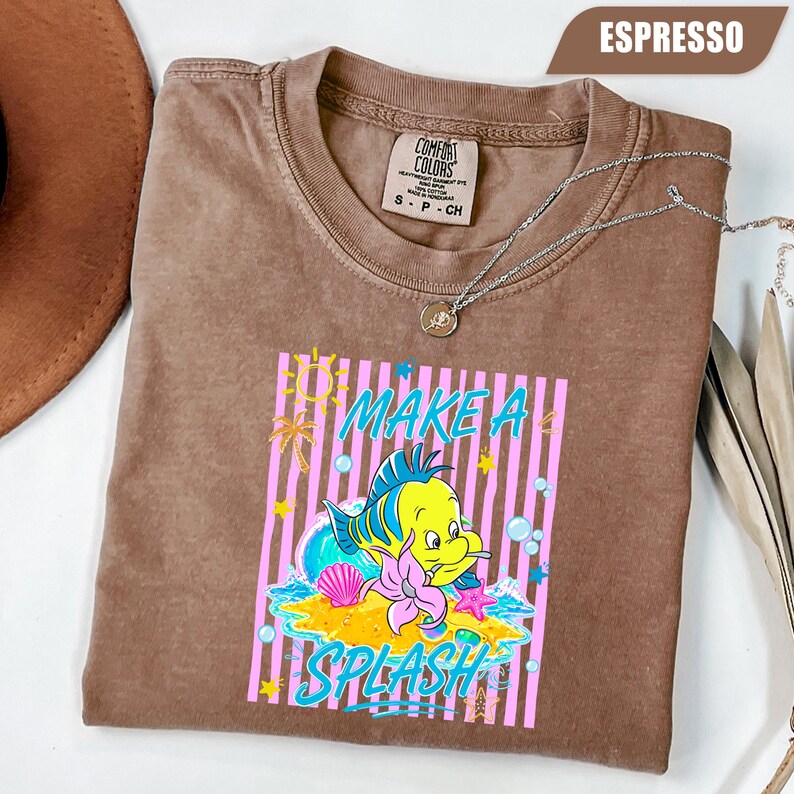 May include: Espresso-colored t-shirt with a graphic of a yellow and blue cartoon fish with the text "MAKE A SPLASH". The design includes pink stripes, a seashell, and a starfish. A silver necklace is draped across the shirt.