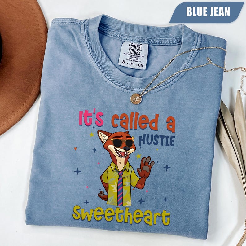 K&ouml;nnte beinhalten: Ein blaues Comfort Colors T-Shirt mit einem Cartoon-Fuchs, der eine Sonnenbrille und eine Krawatte tr&auml;gt. Auf dem Shirt steht in Rosa und Gelb der Text "It's called a HUSTLE Sweetheart". Eine goldene Halskette liegt &uuml;ber dem Shirt.