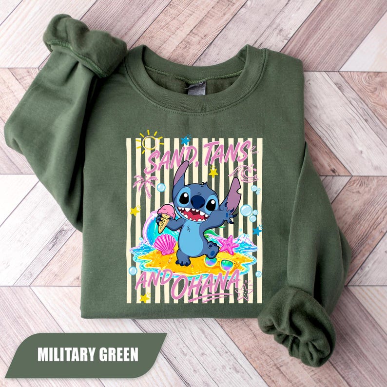 May include: A military green crewneck sweatshirt featuring a graphic of the character Stitch holding an ice cream cone. The design includes the text "SAND, TANS AND OHANA" with beach-themed elements like a seashell, starfish, and sunglasses.