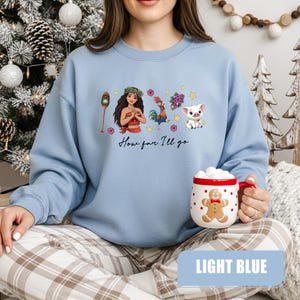 Pu&ograve; includere: Felpa azzurra con una grafica colorata di personaggi dei cartoni animati e la scritta "How far I'll go". &Egrave; visibile una tazza con un omino di pan di zenzero e un bordo rosso.